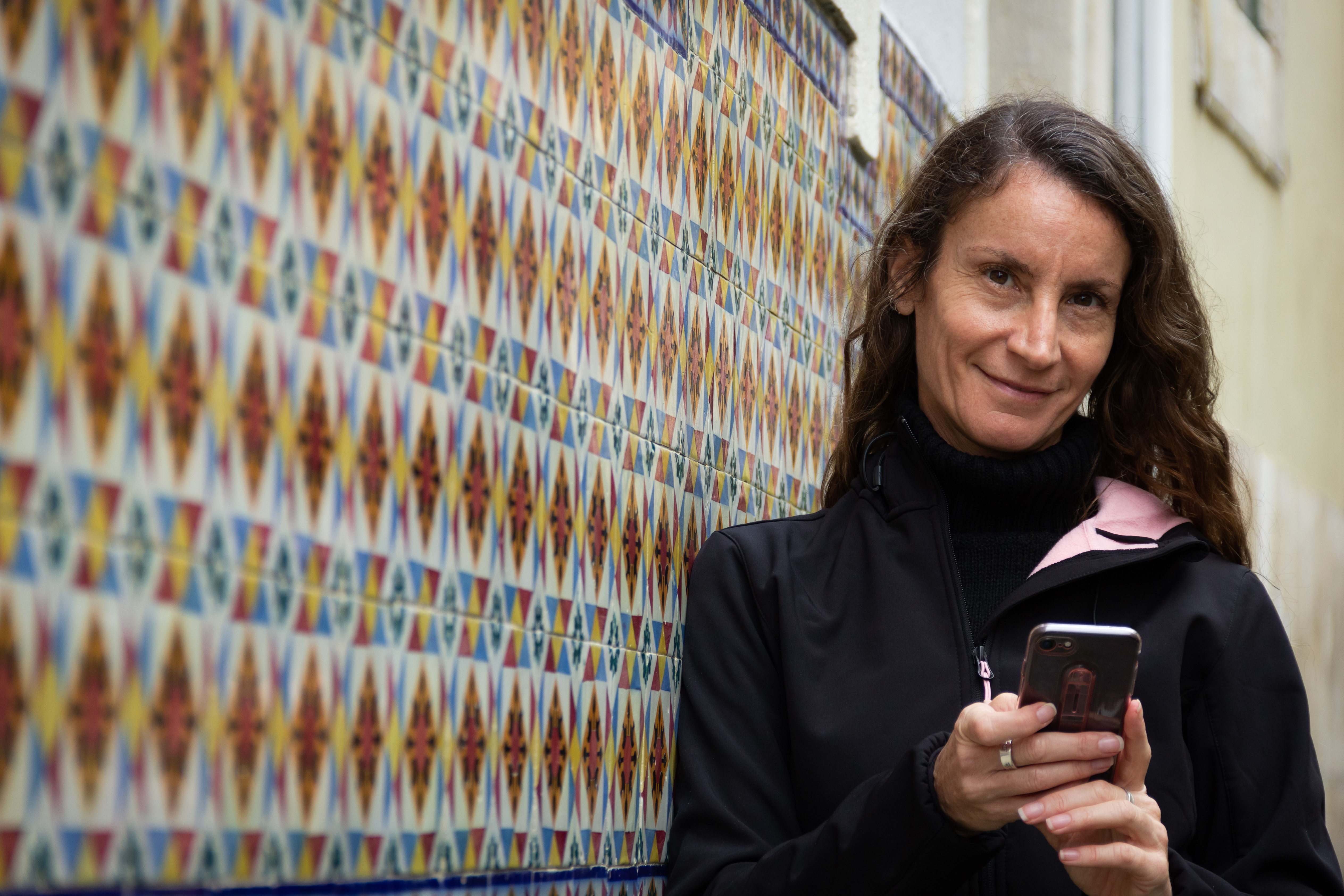 Smiling woman looking at camera holding cellphone and leaning on traditional tile wall building in Lisbon, Portugal. Connection, communication concepts