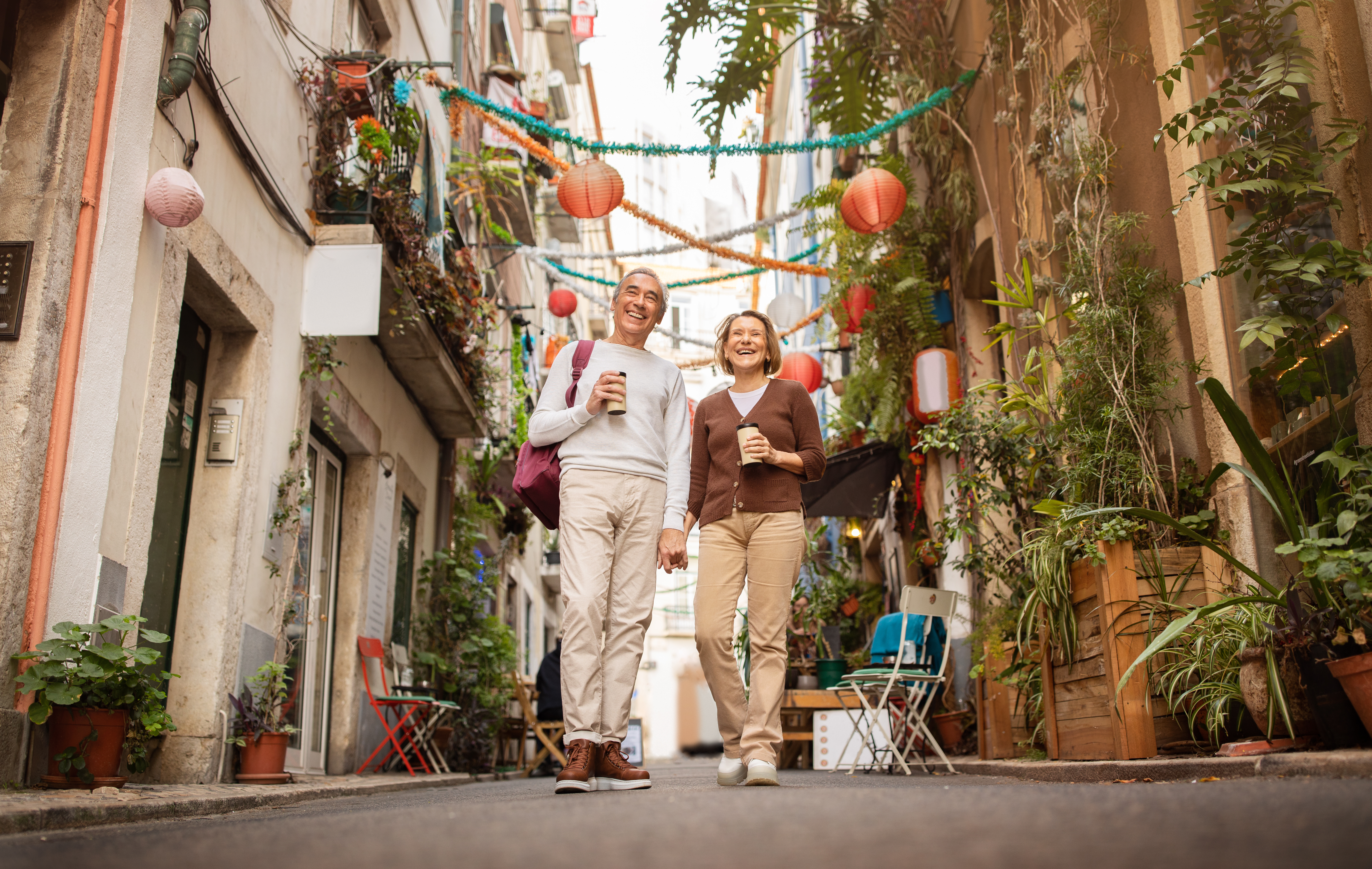 Cheerful Senior Travelers Couple Enjoying Walking Tour Holding Hands And Drinking Coffee On A Street In Lisbon Outside. Tourists Traveling In Europe After Retirement. Low Angle, Full Length Shot