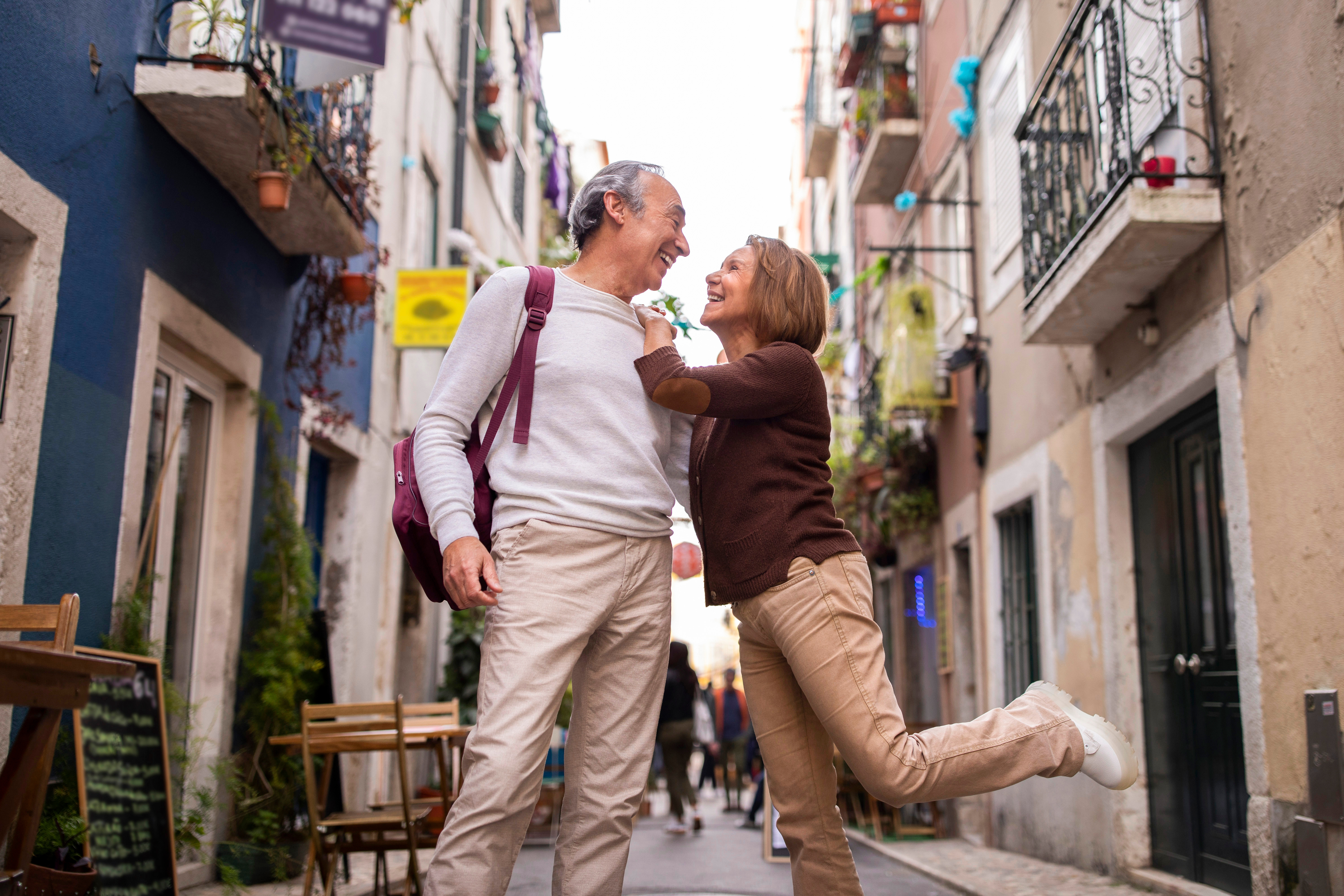 Vacation. Joyful Senior Couple Posing And Having Fun During Walk On A Lisbon Street Outside. Loving Mature Woman Leaning On Husband's Shoulder And Lifting Leg Up. Shot Of Happy Mature Globetrotters